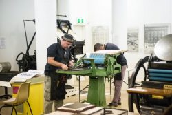 Two men working on an antique proofing press, with linotype machine in background and treadle platen press in foreground