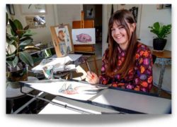 Sami Bayly sitting at drawing table surrounded by illustrations and art supplies, smiling at camera