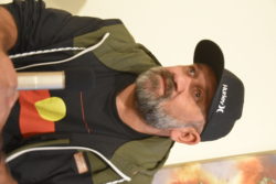 Rob Waters, holding a microphone and wearing an Aboriginal flag T shirt, jacket and black cap, looking off camera