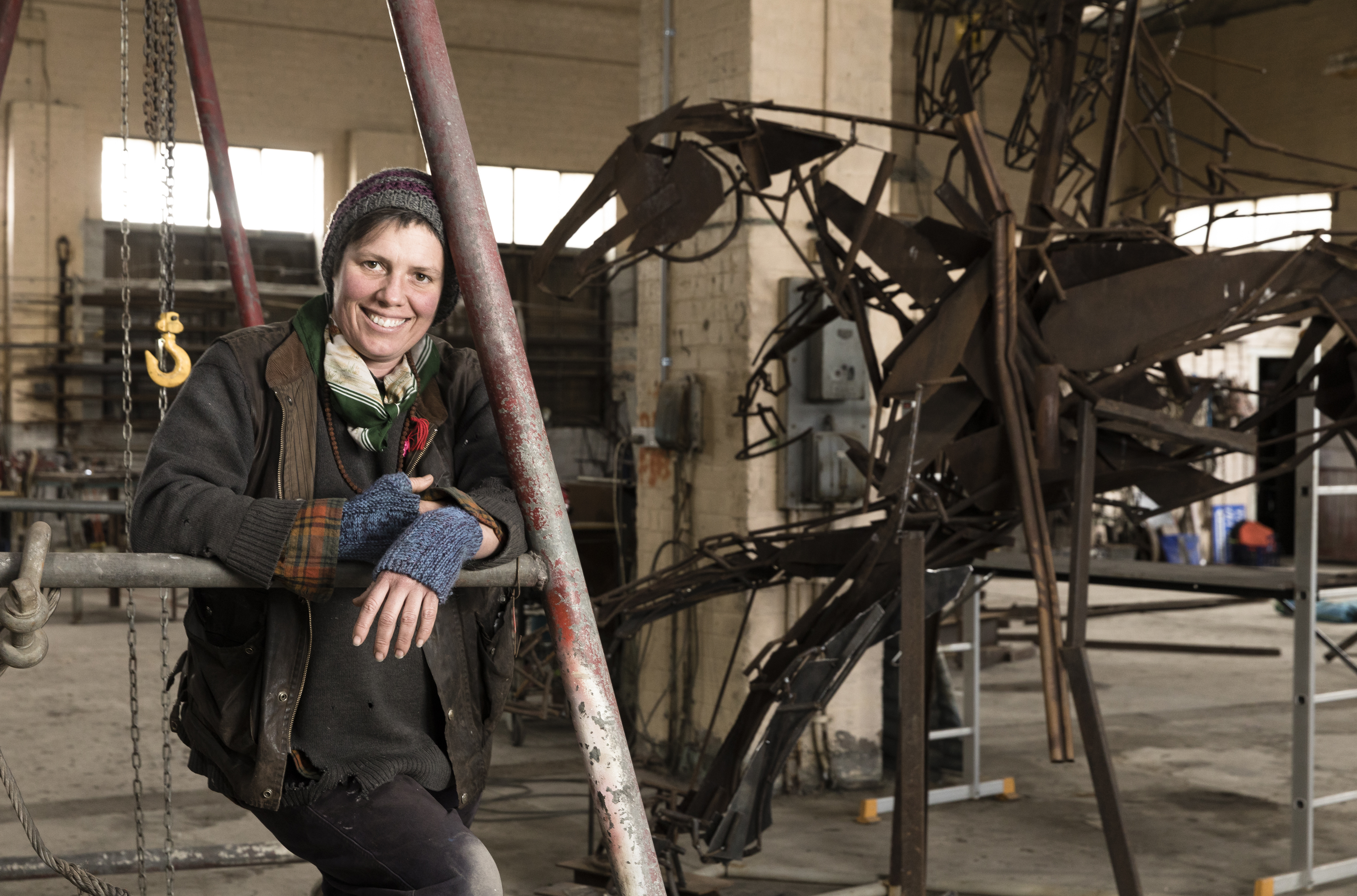Smiling woman in workshop, she is leaning on a metal joist next to a metal horse sculpture