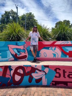 Young man with long hair standing on the whisper wall outside NERAM, featuring a graffiti mural
