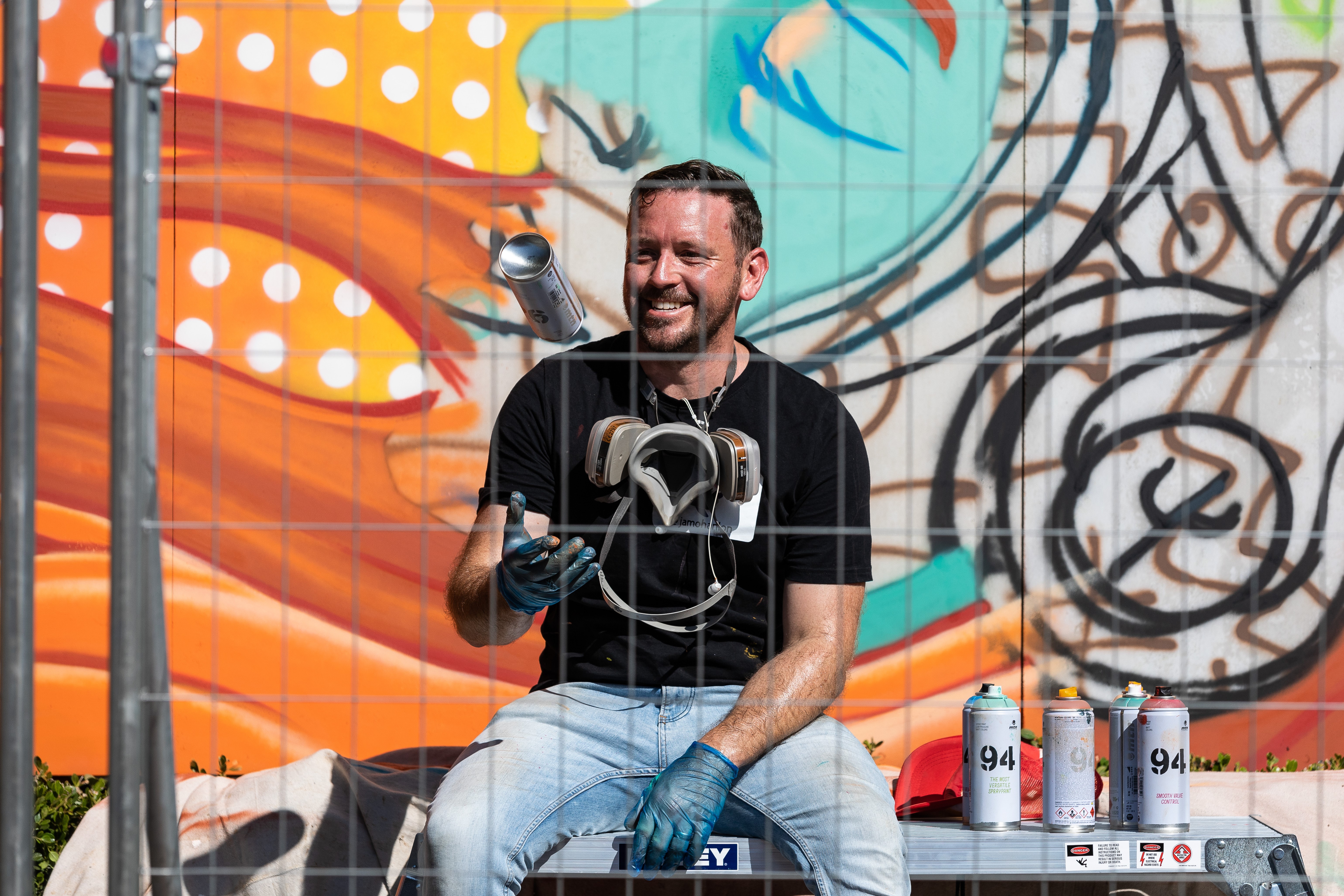Street artist James O'Hanlon sitting in front of a partially finished mural, wearing goves and throwing a can of spray paint in the air