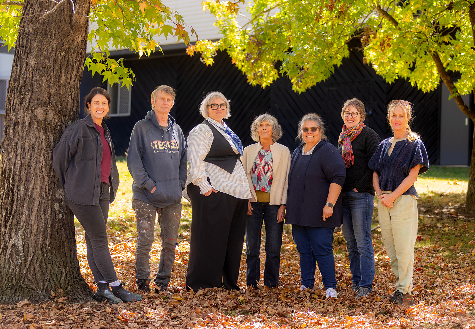 7 people standing beneath a tree with autumn leaves on the ground.